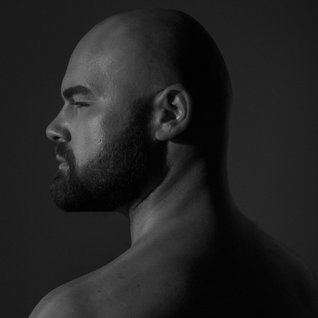 Profile portrait of James Djordjevic, shaved head and dark beard, bare shoulders, against a near-black background.