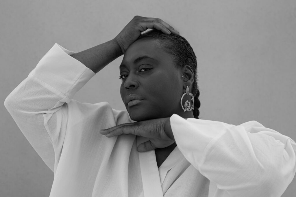 A woman in a white shirt looks straight into the camera, one hand resting on her head, the other tucked under her chin, a large ornate silver earring at her jaw.