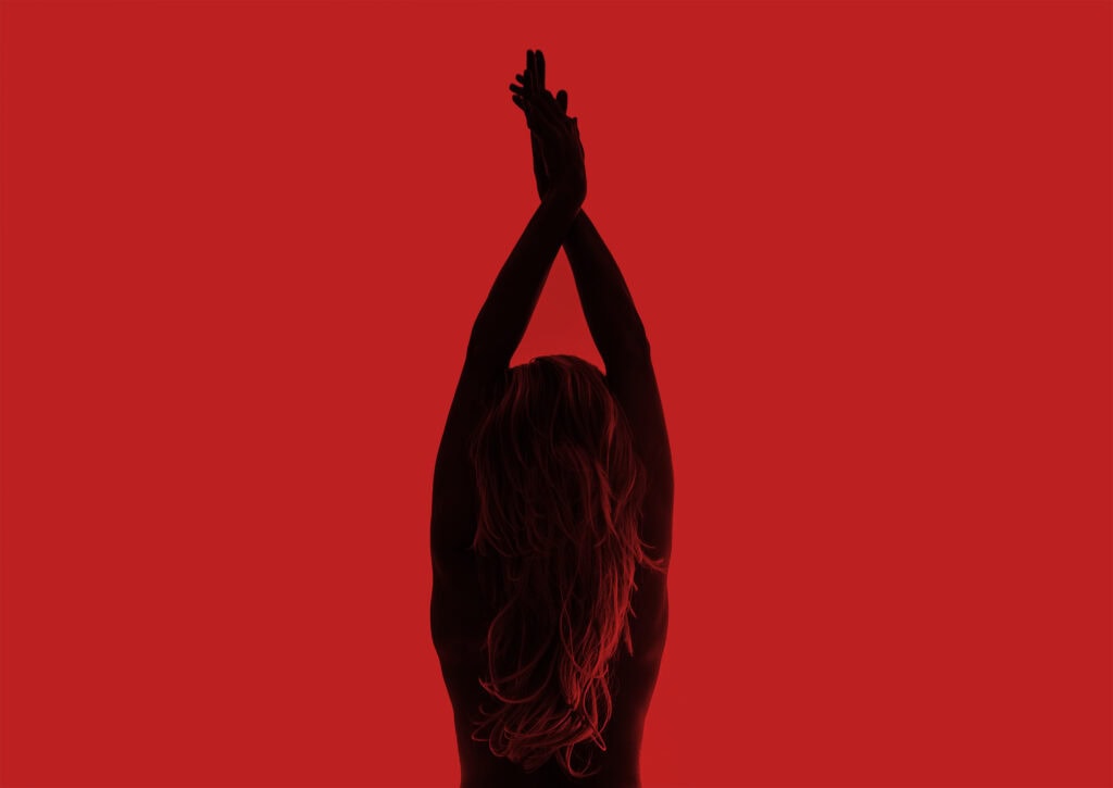 A nude figure in silhouette against a flat red background, arms stretched overhead with wrists together.