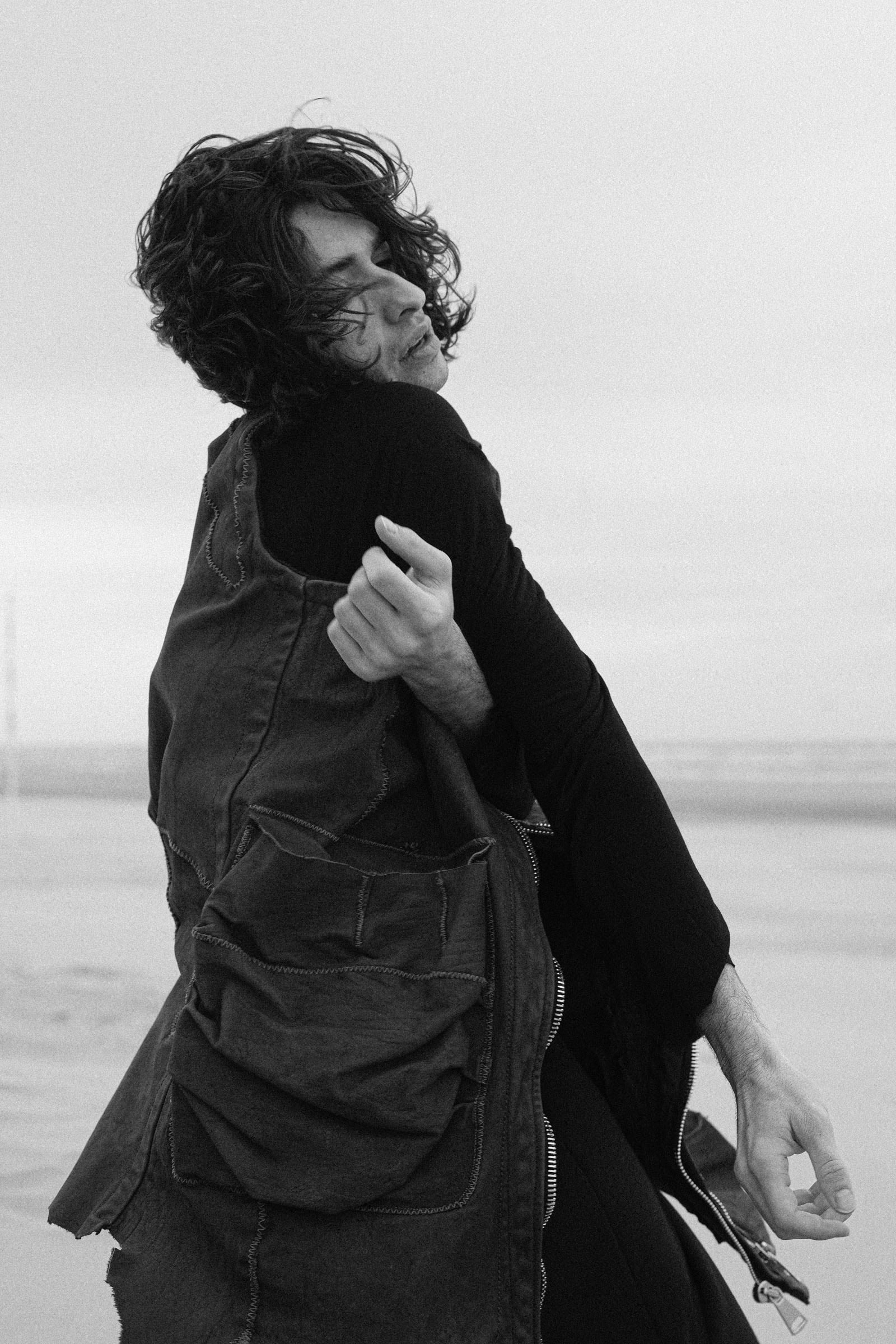 The same man mid-turn on the sand, hair thrown across his face, arms wrapped over his chest as the coat swings out.