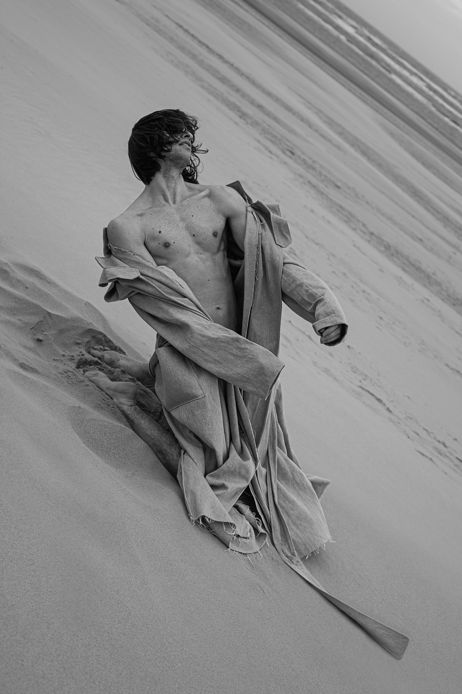 Seen from above, he kneels on a dune with the linen coat sliding off, bare torso, face lifted to the sky.