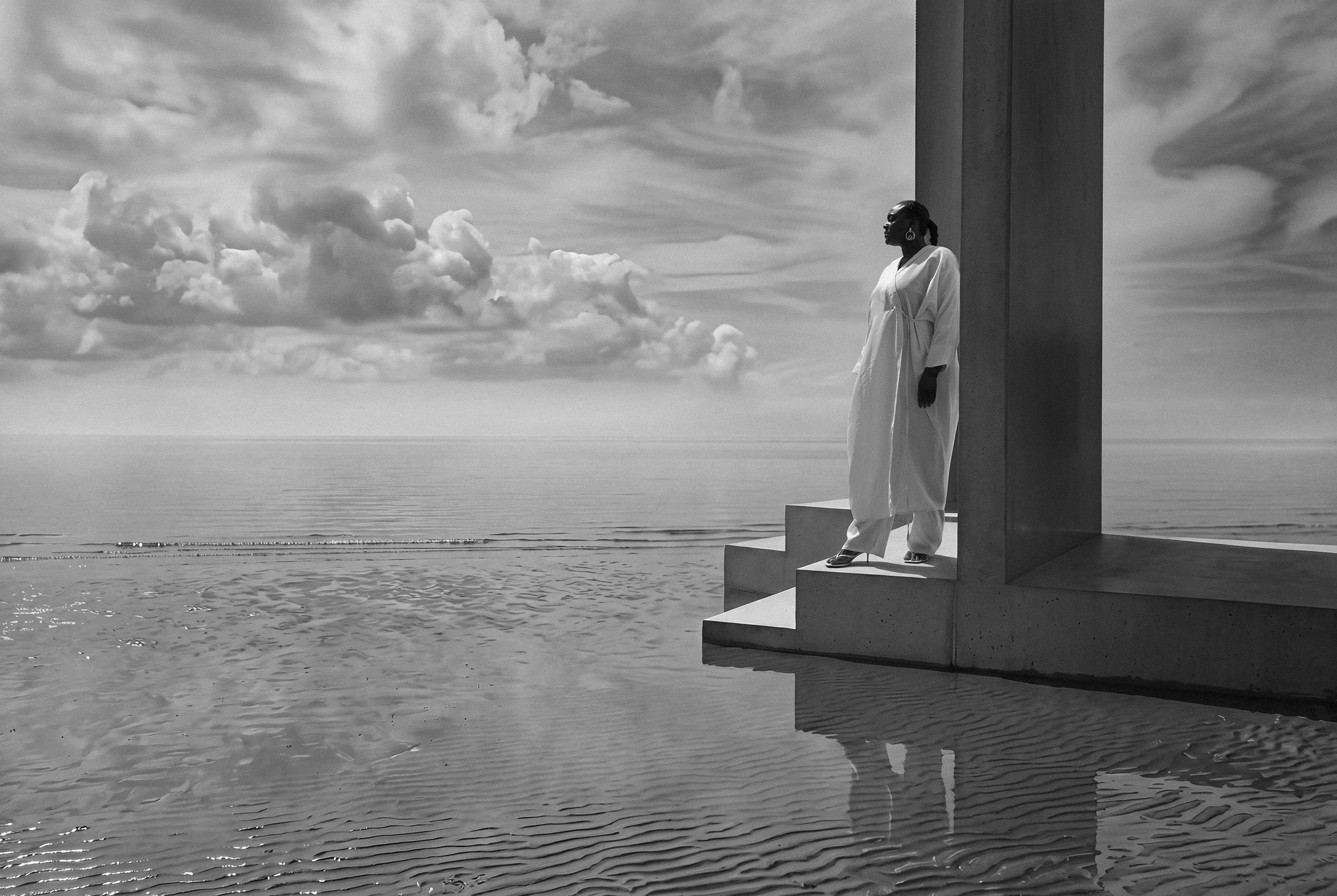 A small white figure stands on concrete steps beside a single tall pillar, a vast plane of still water and towering cloud filling the rest of the frame.